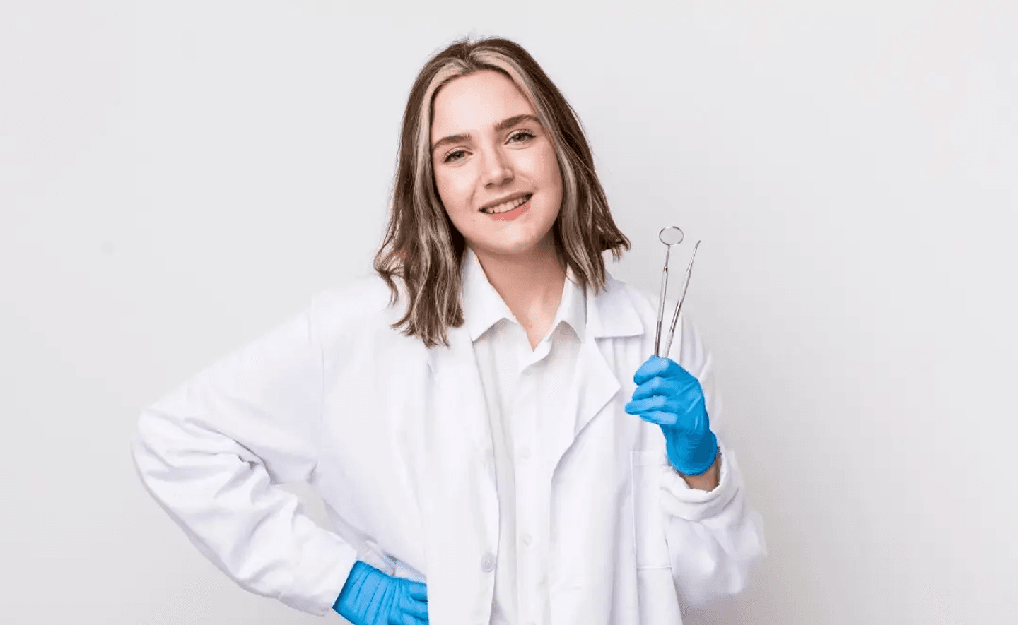 Un profesional de la salud con una bata blanca y guantes azules sostiene instrumentos dentales, de pie frente a un fondo blanco liso.